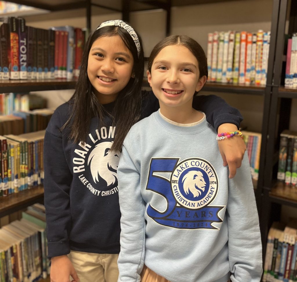 Girls smiling in front of library bookcase, wearing LCCA 50th anniversary and school sweatshirts